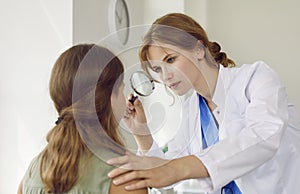 Professional dermatologist examines face of teenage girl with magnifying glass in her office.