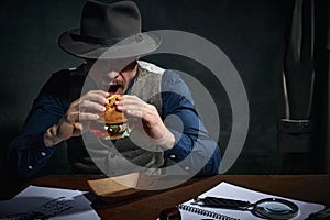 Professional detective in fedora hat sitting at table and eating delicious burger for lunch over dark green vintage