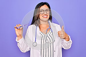 Professional doctor woman showing medical thermometer over purple background happy with big smile doing ok sign, thumb up with
