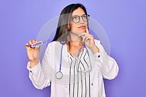 Professional doctor woman showing medical thermometer over purple background serious face thinking about question, very confused