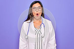 Professional doctor woman wearing stethoscope and medical coat over purple background afraid and shocked with surprise expression,