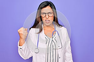 Professional doctor woman wearing stethoscope and medical coat over purple background angry and mad raising fist frustrated and