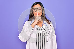 Professional doctor woman wearing stethoscope and medical coat over purple background asking to be quiet with finger on lips