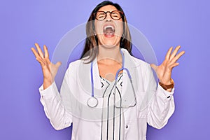 Professional doctor woman wearing stethoscope and medical coat over purple background celebrating mad and crazy for success with