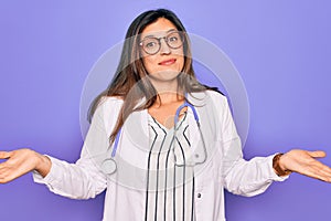 Professional doctor woman wearing stethoscope and medical coat over purple background clueless and confused expression with arms