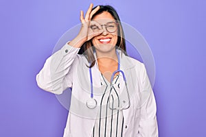 Professional doctor woman wearing stethoscope and medical coat over purple background doing ok gesture with hand smiling, eye