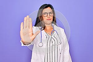 Professional doctor woman wearing stethoscope and medical coat over purple background doing stop sing with palm of the hand