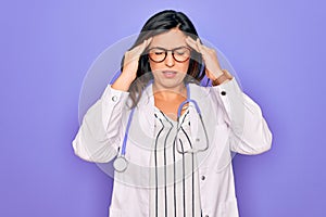 Professional doctor woman wearing stethoscope and medical coat over purple background with hand on head for pain in head because