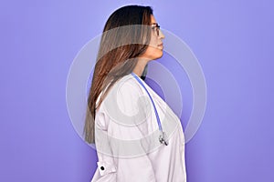 Professional doctor woman wearing stethoscope and medical coat over purple background looking to side, relax profile pose with