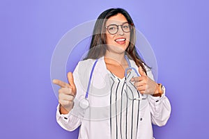 Professional doctor woman wearing stethoscope and medical coat over purple background pointing fingers to camera with happy and