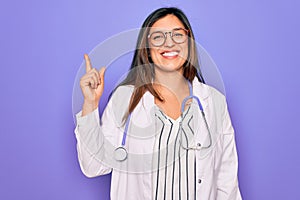 Professional doctor woman wearing stethoscope and medical coat over purple background showing and pointing up with finger number