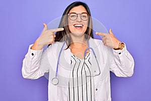 Professional doctor woman wearing stethoscope and medical coat over purple background smiling cheerful showing and pointing with