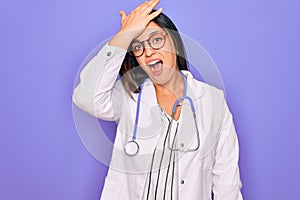 Professional doctor woman wearing stethoscope and medical coat over purple background surprised with hand on head for mistake,