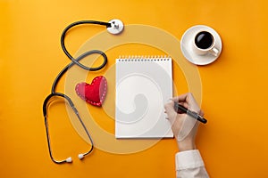 Professional doctor writing medical records in a notebook with stethoscope, coffee cup, syringe and heart