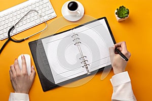 Professional doctor writing medical records in a notebook with stethoscope, keyboard, coffee cup and mouse