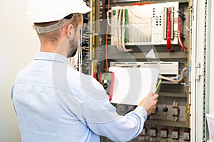 Professional electrician in hardhat reads electric scheme near control panel on substation