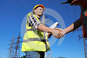 Professional electricians shaking hands near high voltage tower