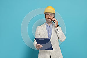 Professional engineer in hard hat with clipboard talking on phone against light blue background