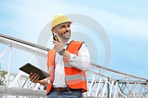 Professional engineer with tablet and walkie talkie near high voltage tower construction outdoors. Installation of electrical