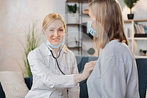 Professional female doctor or nurse doing the auscultation of her senior lady patient at home, listening the heart beat