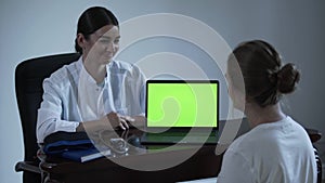 Professional female doctor tells something to female patient and showing with hand on green screen of laptop. Concept of