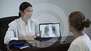 Professional female doctor tells something to female patient and showing on screen of laptop with lung x-ray image