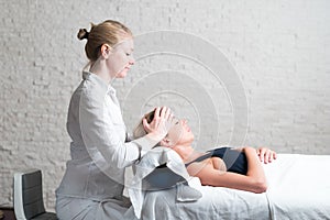 Professional female masseur giving relaxing massage treatment to young female client. Hands of masseuse on forehead of