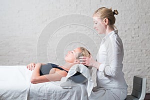 Professional female masseur giving relaxing massage treatment to young female client. Hands of masseuse on forehead of