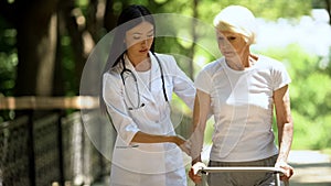 Professional female nurse supporting old woman moving in park with walking frame