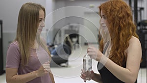 Professional female trainer giving bottle of water to the redhaired sportswoman in the gym. The girl working with