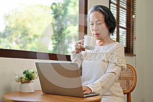 Focused aged Asian businesswoman working from home, sipping coffee while using laptop