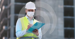 Professional foreman in protective mask and helmet checking building with plan at construction site