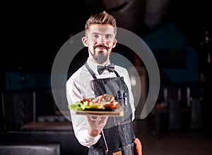 Professional friendly waiter helpfully serves plate with prepared dish on a black background.