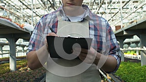 Professional gardener in apron writing notes on clipboard working in greenhouse