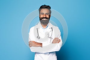 Professional general practitioner. Portrait of cheerful indian male doctor posing with folded arms and smiling at camera