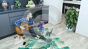 Professional guitarist tuning guitar sitting on floor in kitchen at home. Musician tunes guitar staying at home during quarantine.