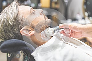 Professional hairdresser applying shaving foam on client skin in barbershop