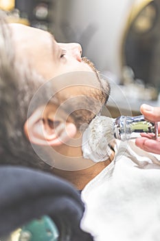 Professional hairdresser applying shaving foam on client skin in barbershop