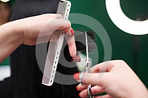 Professional hairdresser making haircut for female client. Close-up picture of hair stylist`s hands holding scissors and comb,