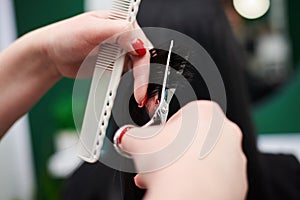 Professional hairdresser making haircut for female client. Close-up picture of hair stylist`s hands holding scissors and comb,