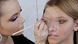 A professional make-up artist applies a special brush with eyeliner in beige color to the eyes of a young beautiful girl