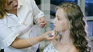 Professional make-up artist applying tonal cream to blond long curl hair and blue eyes model skin.