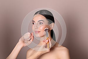 Professional makeup artist applying powder onto beautiful young woman`s face with brush on dusty rose background