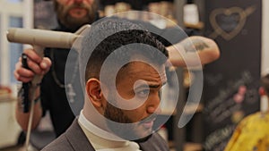 Professional male barber arranges his indian client's hair. He uses a brush and a dryer for this.