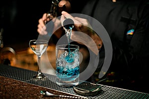 Professional male bartender pouring a blue alcoholic drink from the jigger to a measuring cup holding a glass bottle