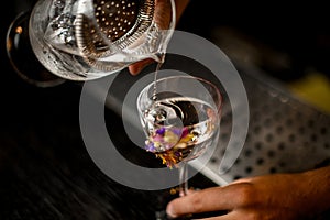 Professional male bartender pouring a cocktail from the measuring cup with a strainer to a glass decorated with flowers