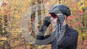 Professional male cyclist in black sports apparel and helmet preparing for training in autumn park and puts on glasses. Cycling tr