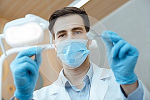 Professional male dentist in white coat, protective mask and gloves hold dental instruments and start job