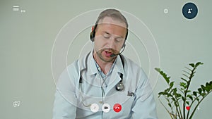 Professional male doctor in white medical coat and headset making conference call on laptop computer, consulting