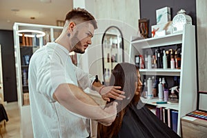 Professional male hairdresser brushing long brown hair of female client, making hairstyle at beauty salon